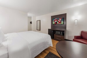 A hotel room with a bed, a TV mounted on the wall, a small kitchenette, and a seating area with a table and chair.