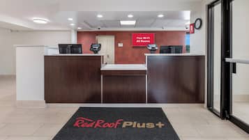 A reception area with a Red Roof Plus sign, two reception desks, a clock, and a sign indicating free Wi-Fi in all rooms.