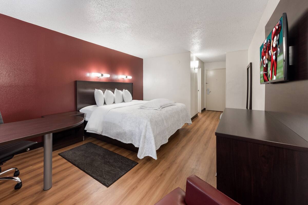 A modern hotel room with a neatly made bed, a desk with a chair, and a flat-screen TV mounted on the wall.