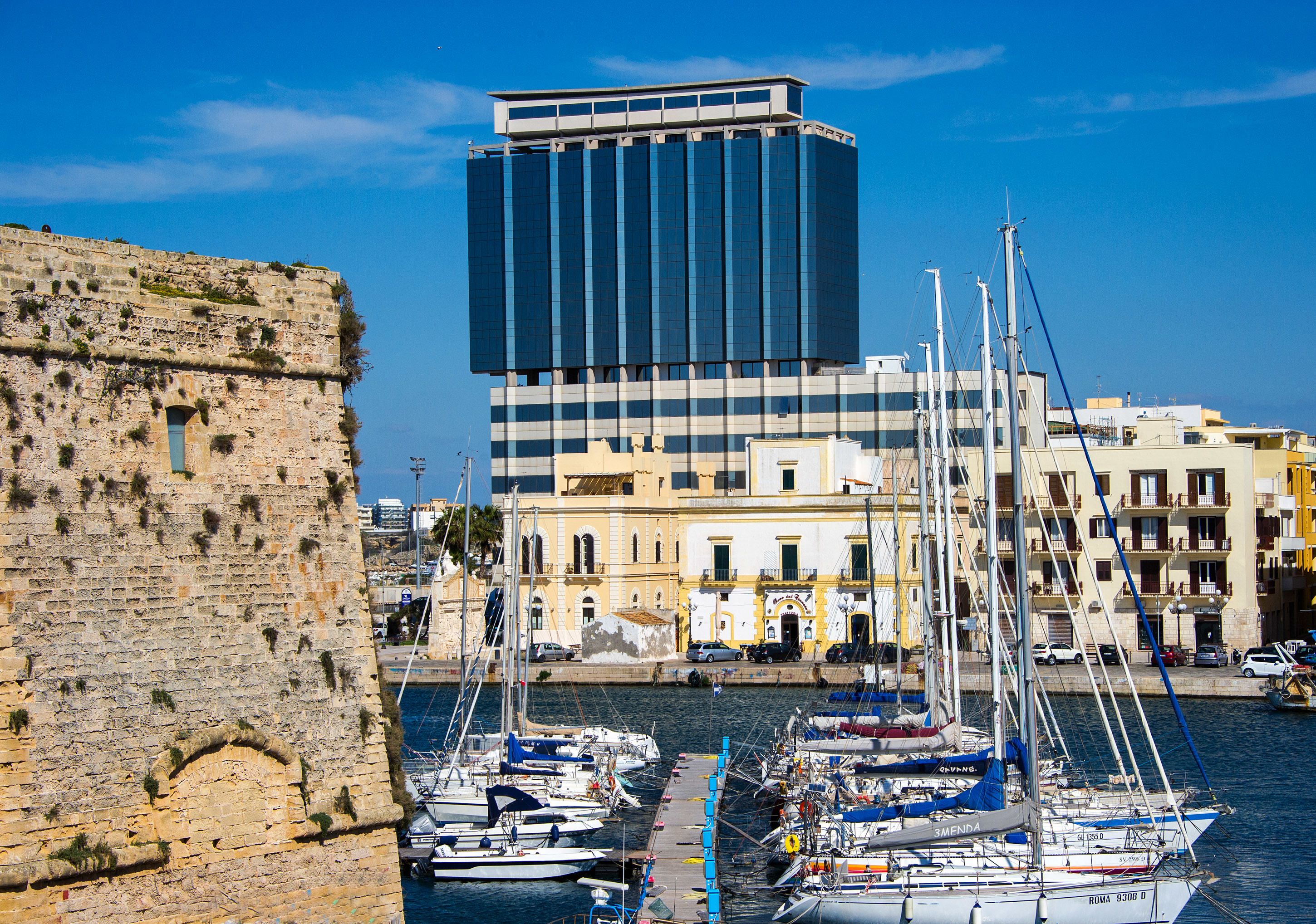 A harbor with multiple sailboats docked, a historic stone wall, and a modern building in the background.
