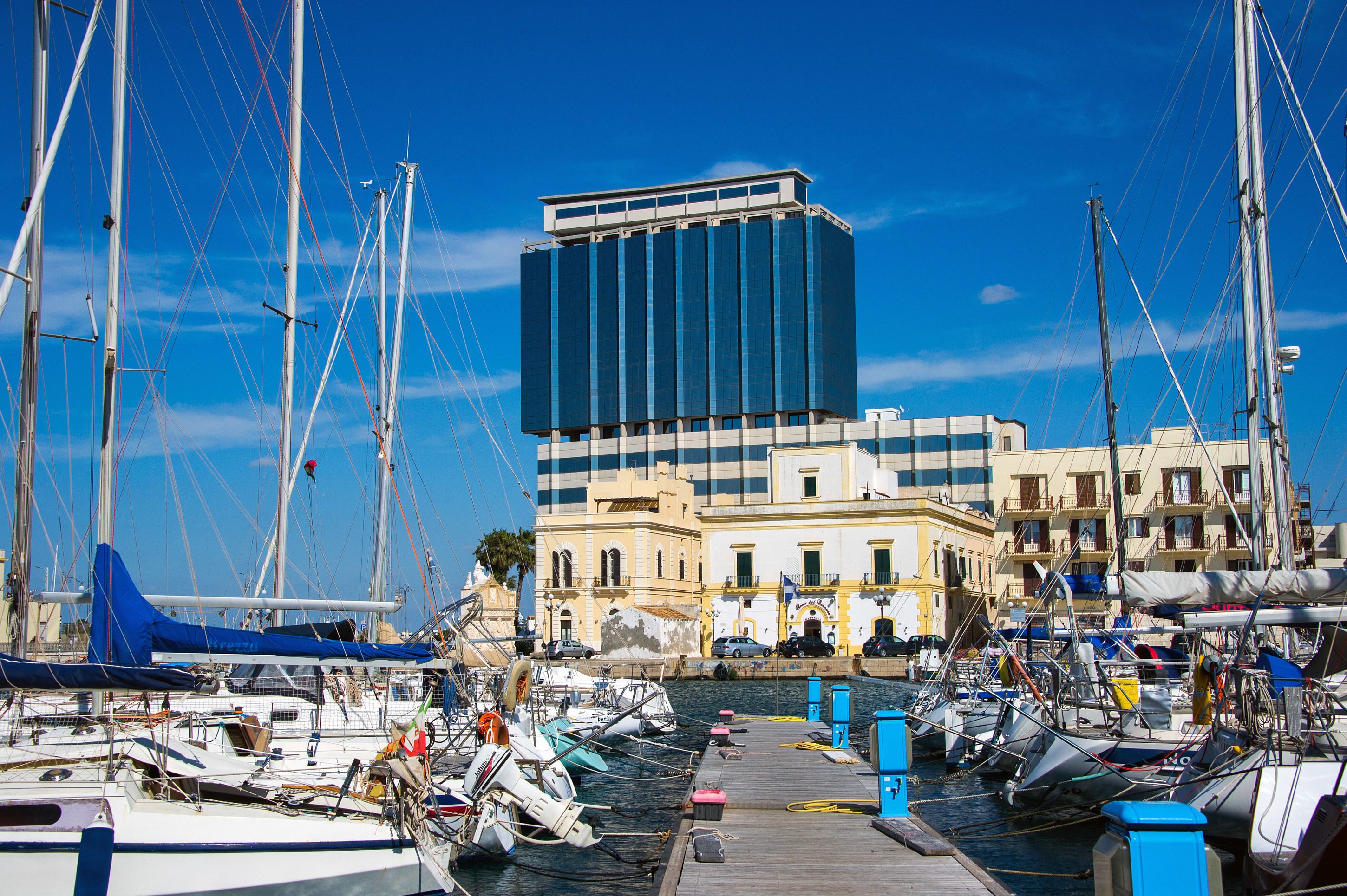 A marina with multiple boats docked, a modern building with a blue facade, and traditional architecture in the background.