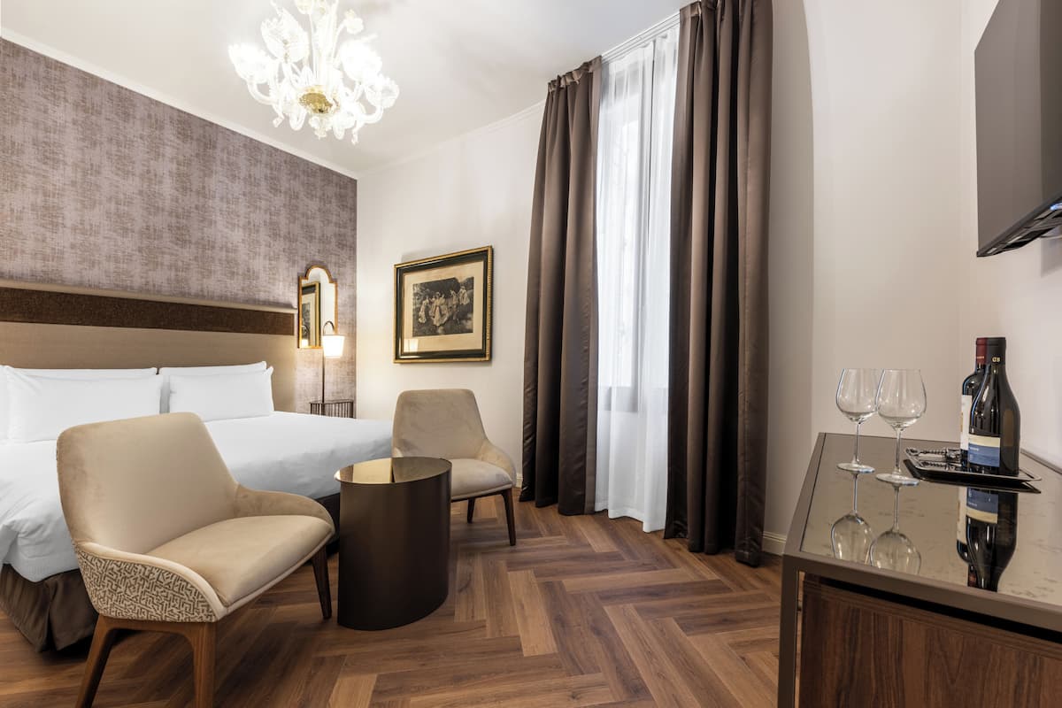 Habitación de hotel con cama, dos sillas, una mesa redonda, un espejo, un cuadro en la pared y un minibar con copas de vino.