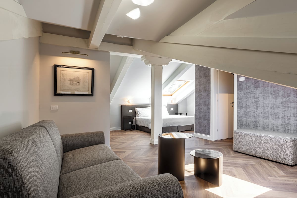 Una habitación de hotel moderna con una cama de altillo, un sofá gris, una mesita redonda y un cuadro en la pared.