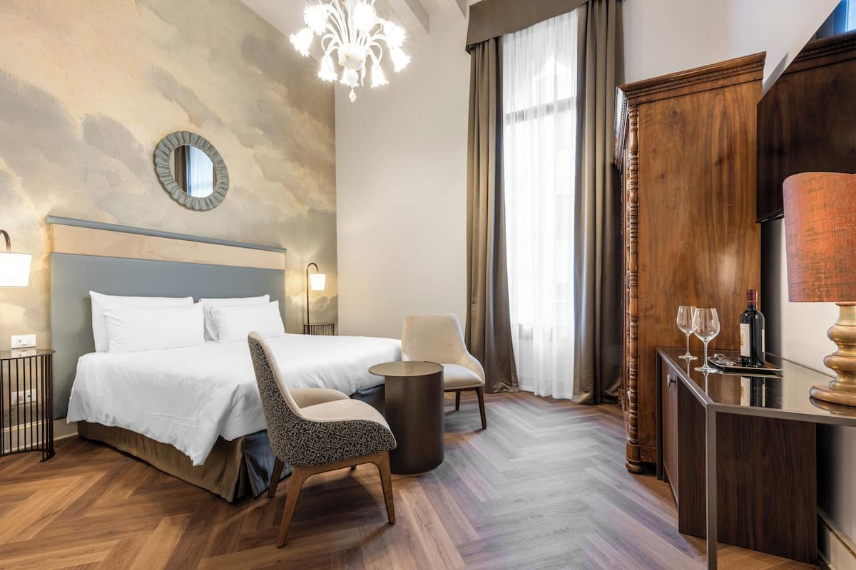 Habitación de hotel con cama, dos sillones, una mesita con vasos y una botella, un espejo redondo y una araña.