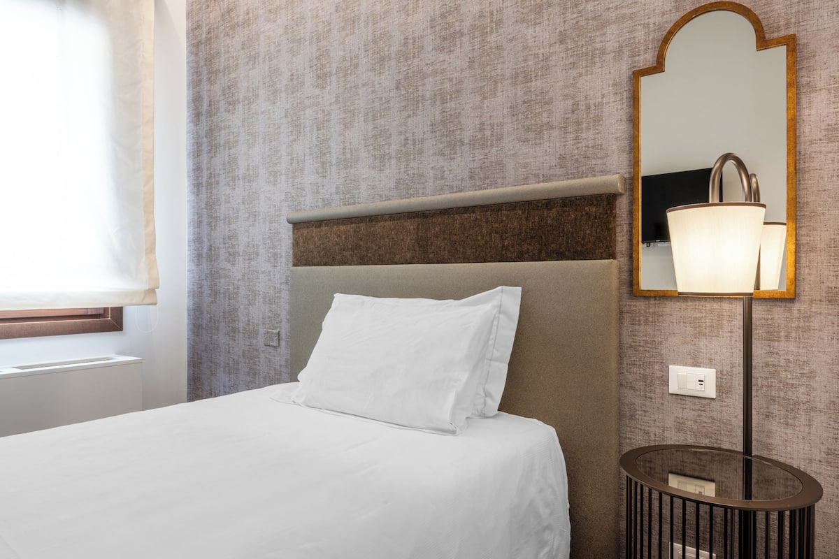 Habitación de hotel con cama, mesita de noche, una mesa redonda, espejo en la pared y ventana con cortinas.