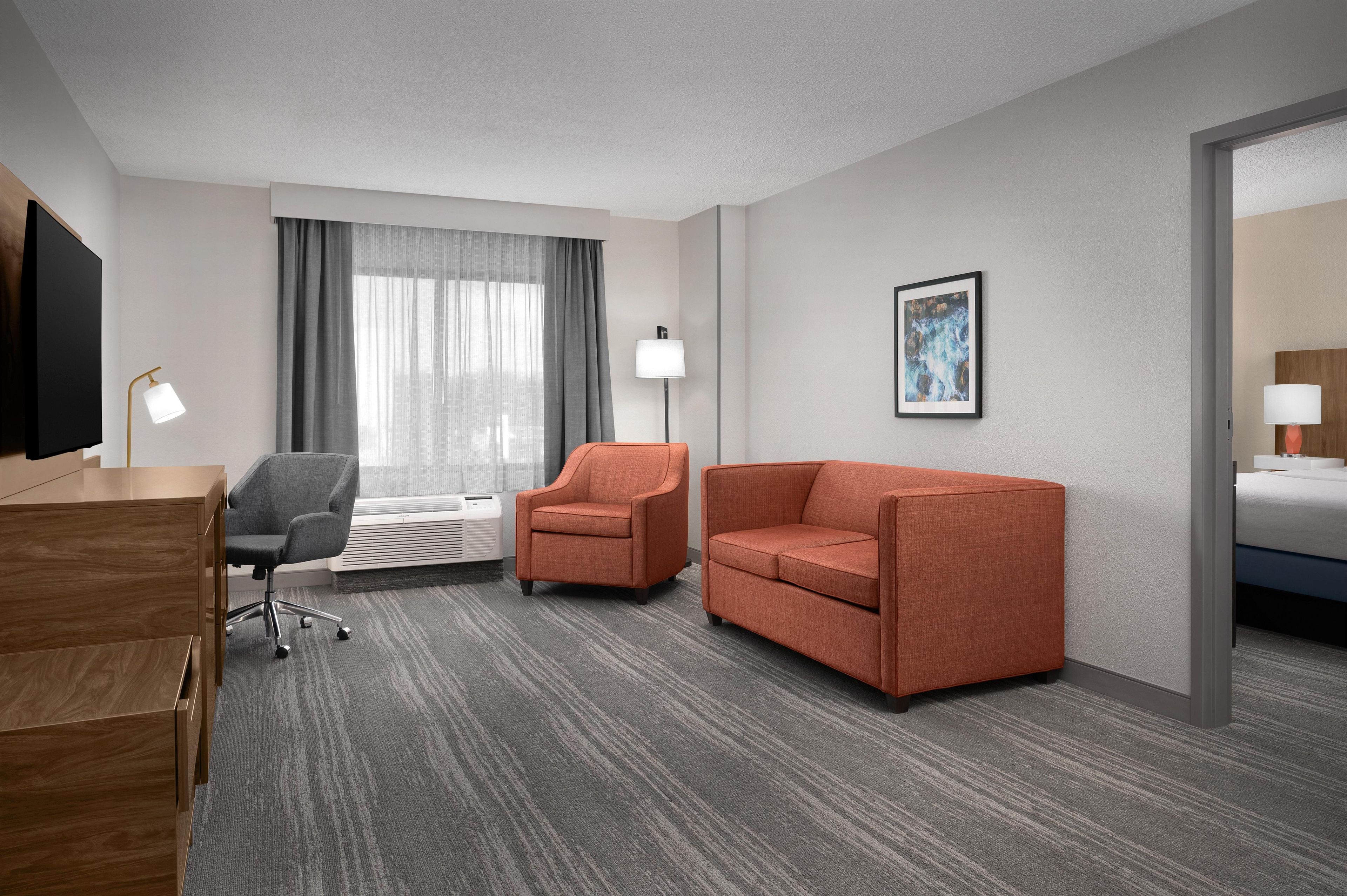 Habitación de hotel con escritorio, dos sillones, un sofá, una cama, ventana con cortinas, televisor de pared, cuadro enmarcado y lámpara.
