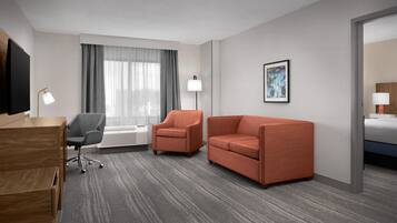 Habitación de hotel con escritorio, dos sillones, un sofá, una cama, ventana con cortinas, televisor de pared, cuadro enmarcado y lámpara.