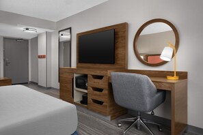 A hotel room with a bed, a wooden desk with a flat-screen TV, a mirror, a desk lamp, and a grey office chair. - Hampton Inn & Suites Denver - Cherry Creek (Glendale)