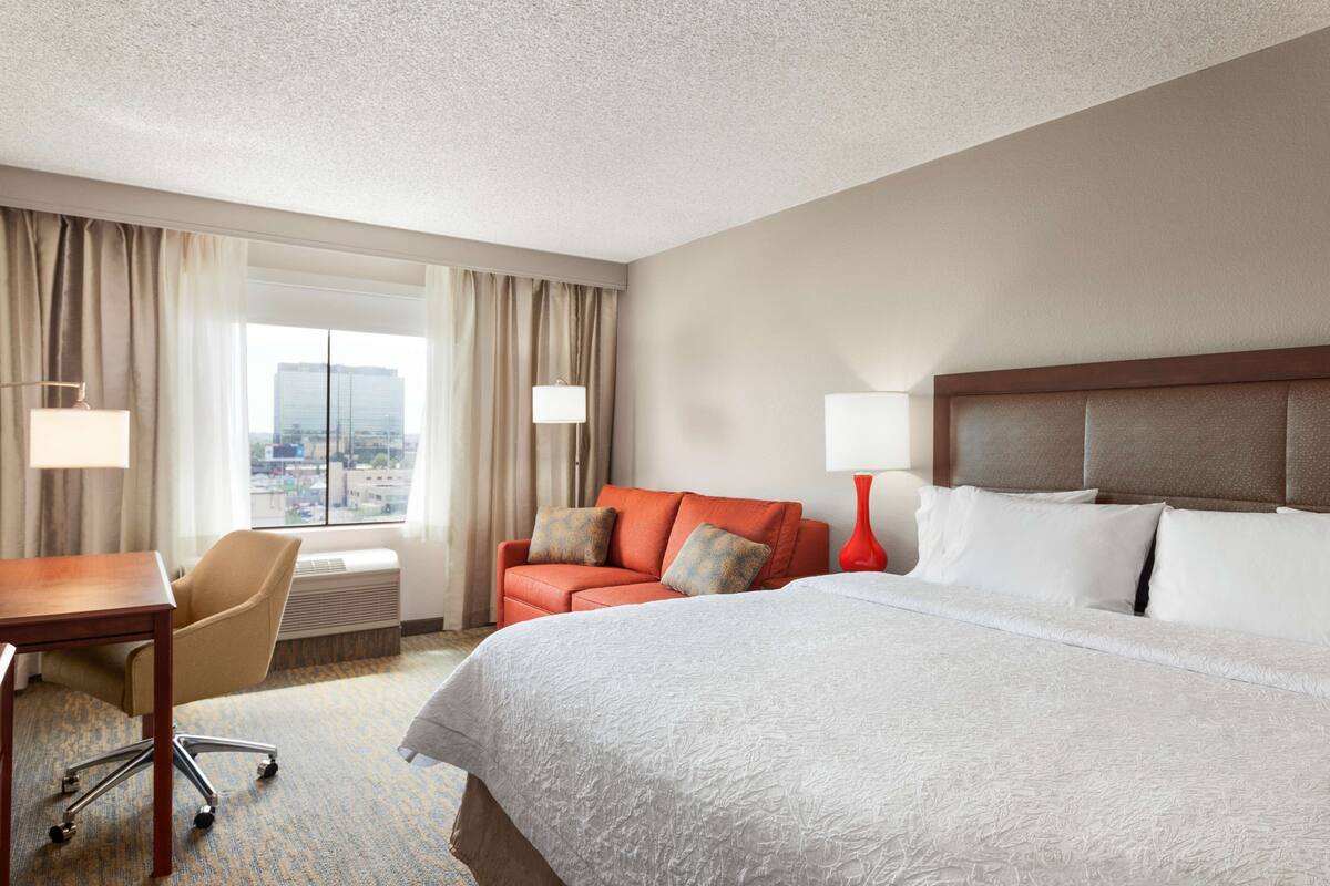 A hotel room with a large bed, a desk, a chair, and a view of the city through the window.