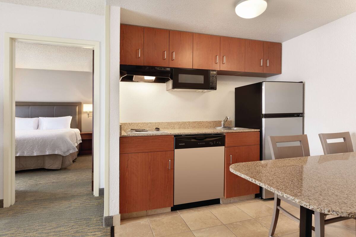 A compact hotel room with a kitchenette, a bed with white linens, and a dining area with a table and chairs.