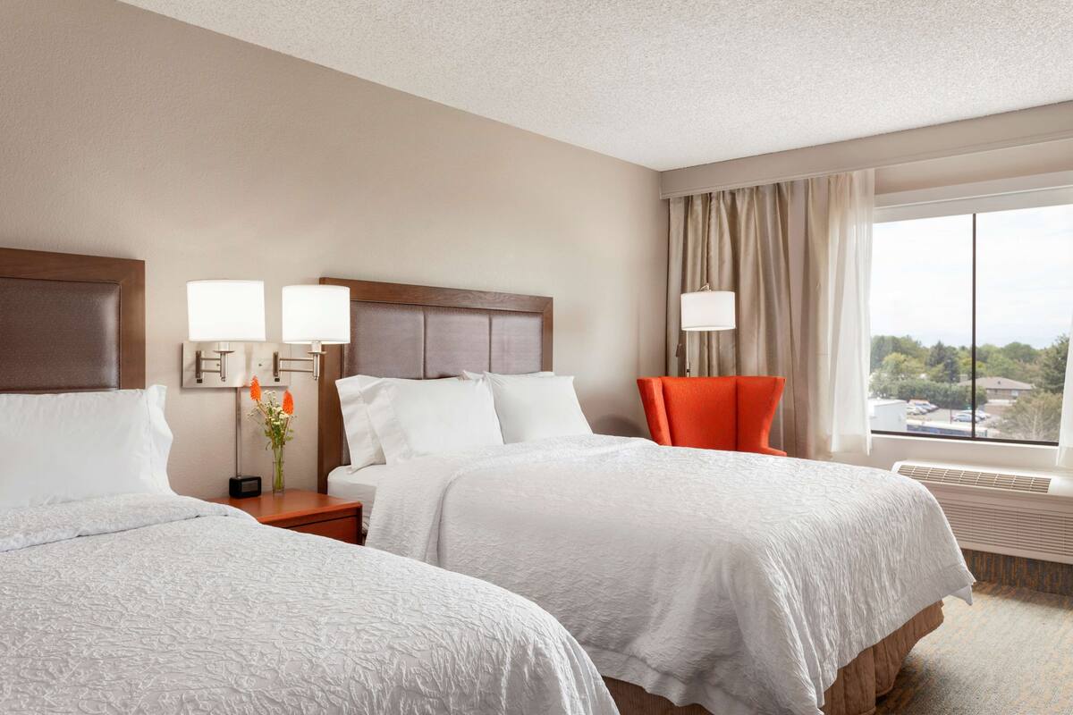 A hotel room with two beds, a nightstand with lamps, a vase with flowers, a chair, and a large window with a view.