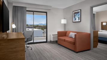 A hotel room with a sofa, a desk, a chair, a bed, a TV, a lamp, and a painting on the wall.