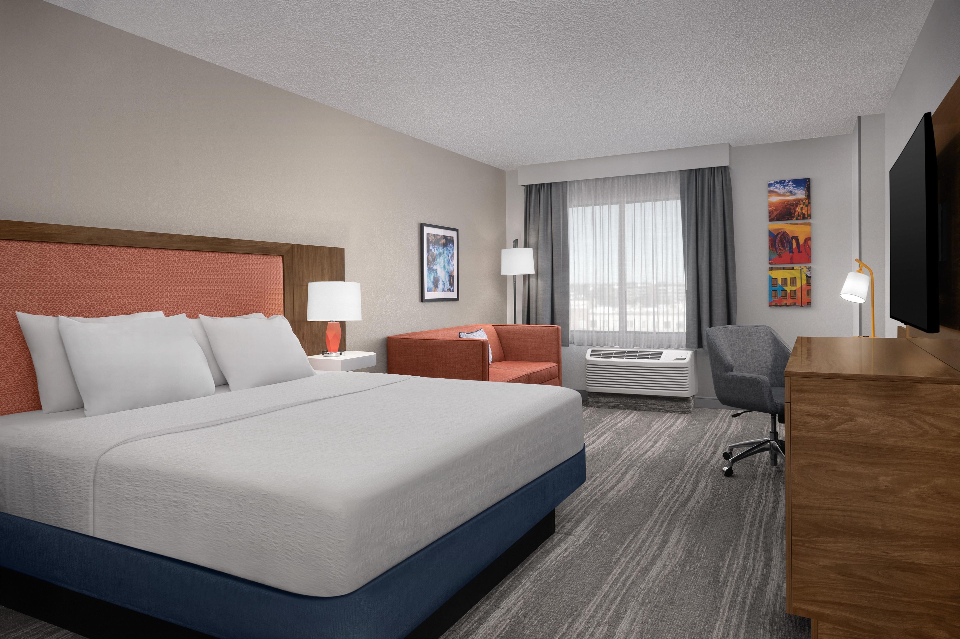 A hotel room with a large bed, a desk, a chair, a TV, and an air conditioning unit.
