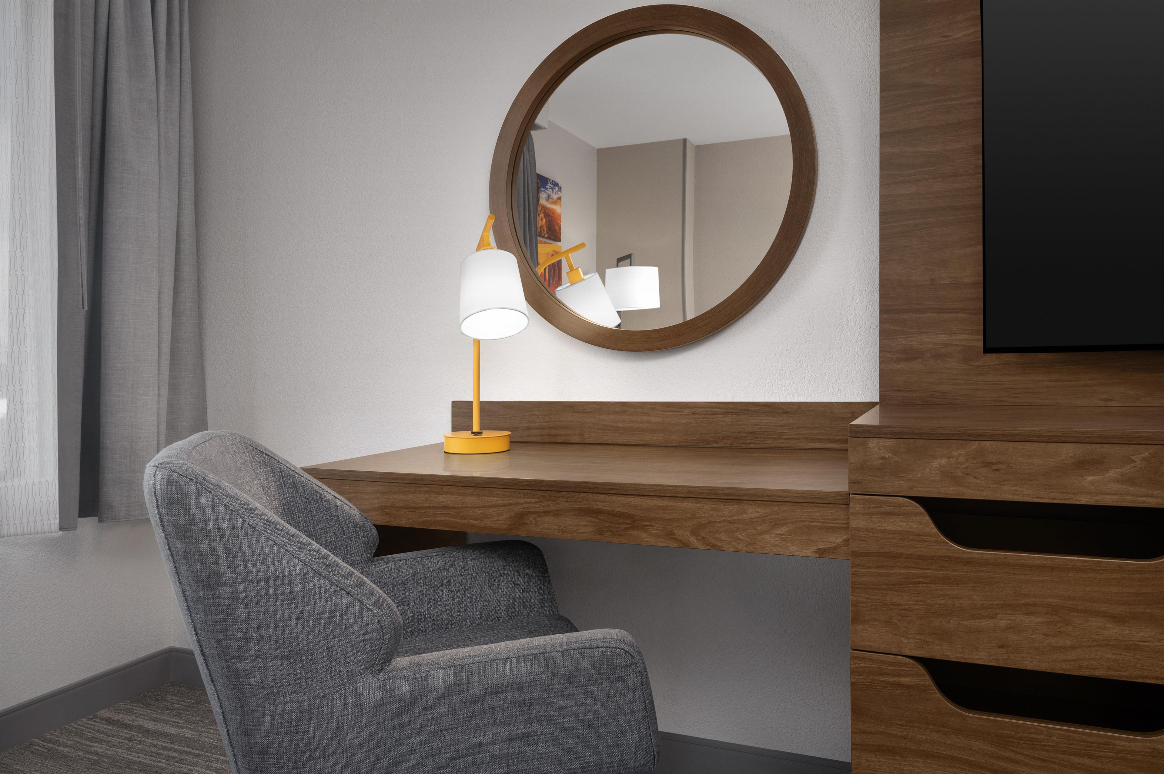 A modern hotel room with a grey armchair, a wooden desk with a mirror, and a yellow desk lamp.