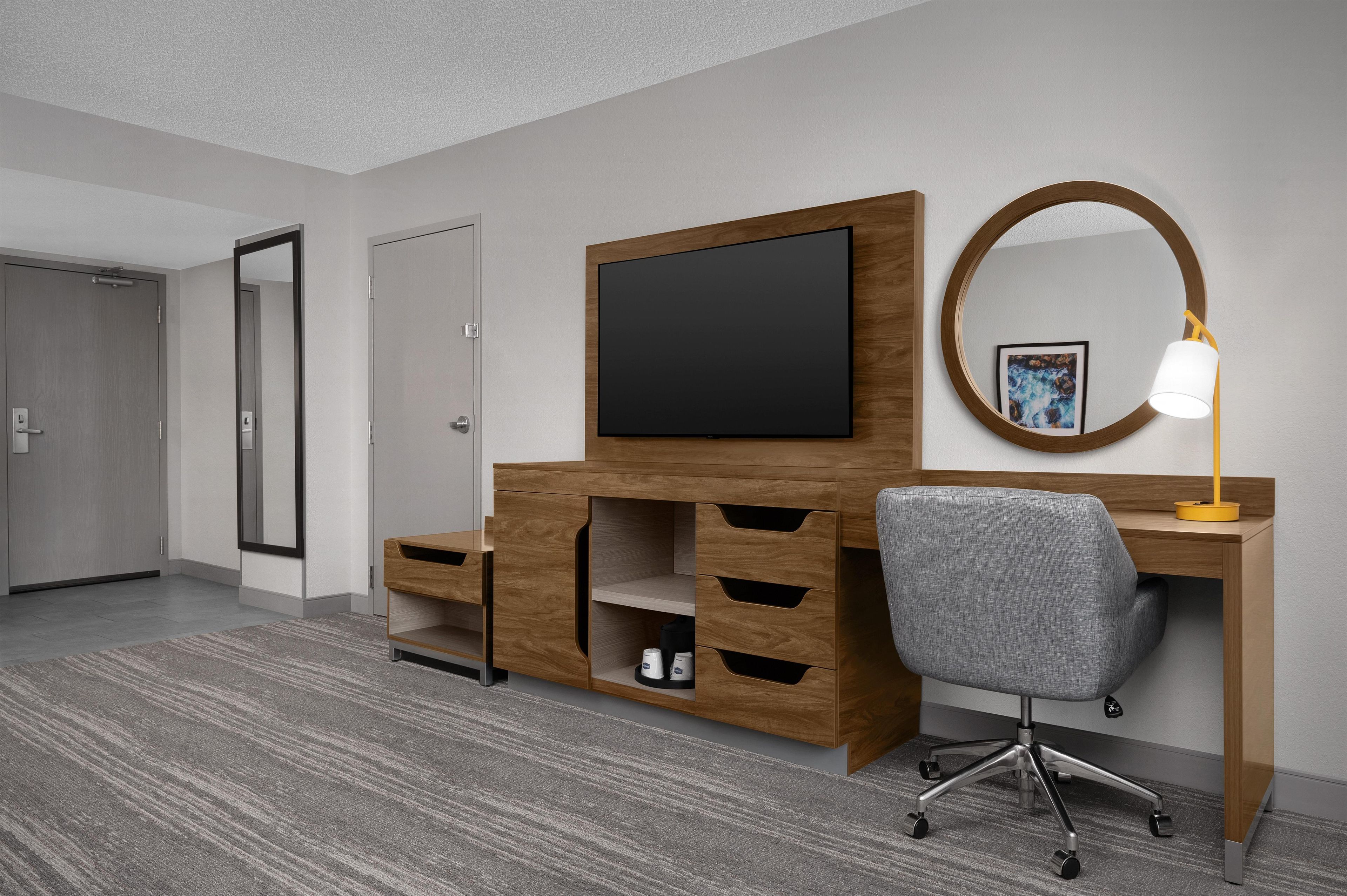 A hotel room with a wooden desk, a grey office chair, a flat-screen TV, a bedside lamp, and a large mirror.