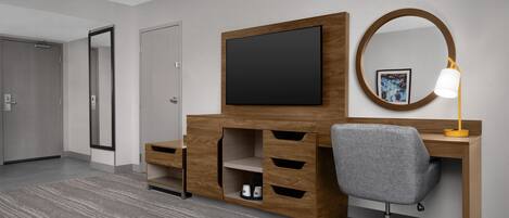 A hotel room with a wooden desk, a grey office chair, a flat-screen TV, a bedside lamp, and a large mirror.