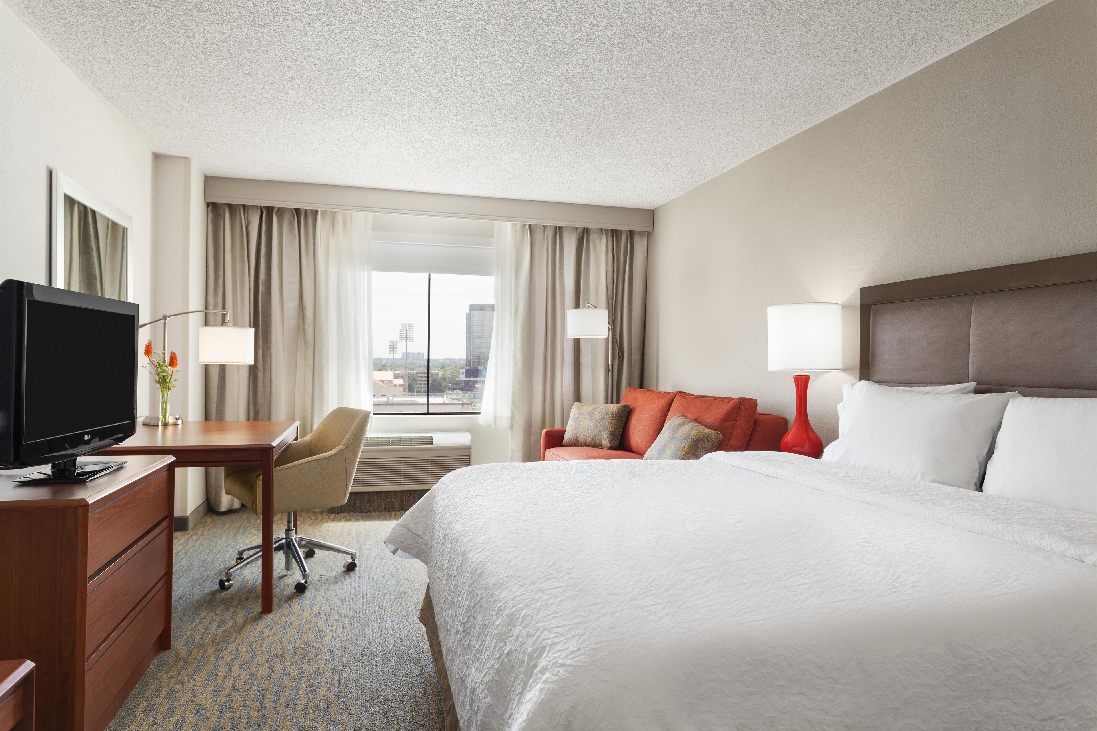 A hotel room with a bed, a desk with a TV, a chair, a window with curtains, and a city view.
