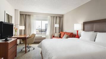 A hotel room with a bed, a desk with a TV, a chair, a window with curtains, and a city view.