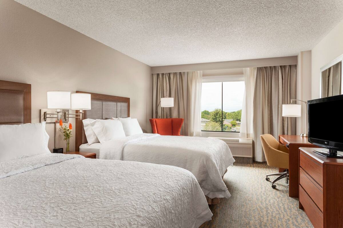 A hotel room with two beds, a desk, a TV, and a view of the outdoors.