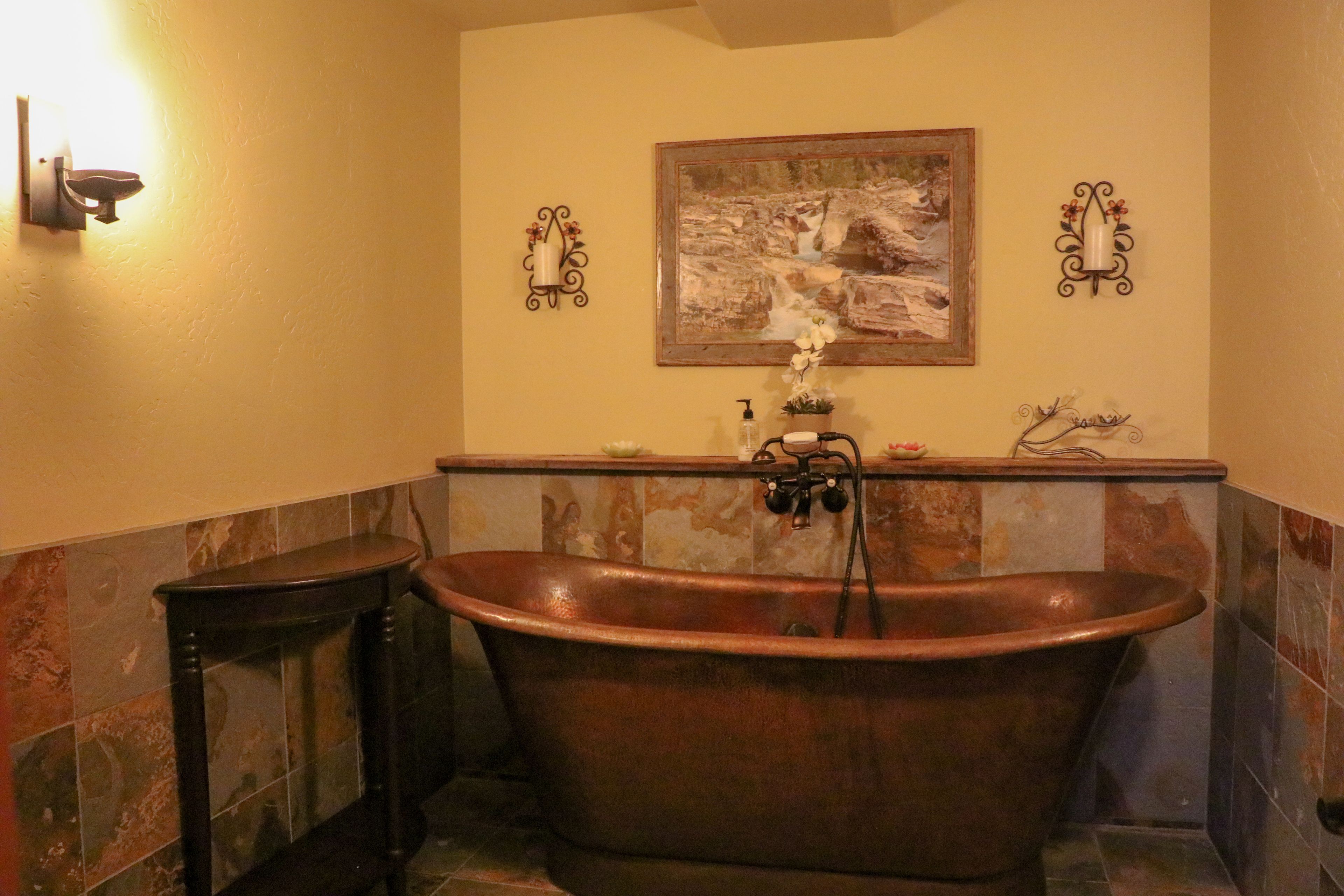 Une salle de bain avec une baignoire sur pieds, une petite table, un luminaire fixé au mur et un tableau encadré au-dessus de la baignoire.