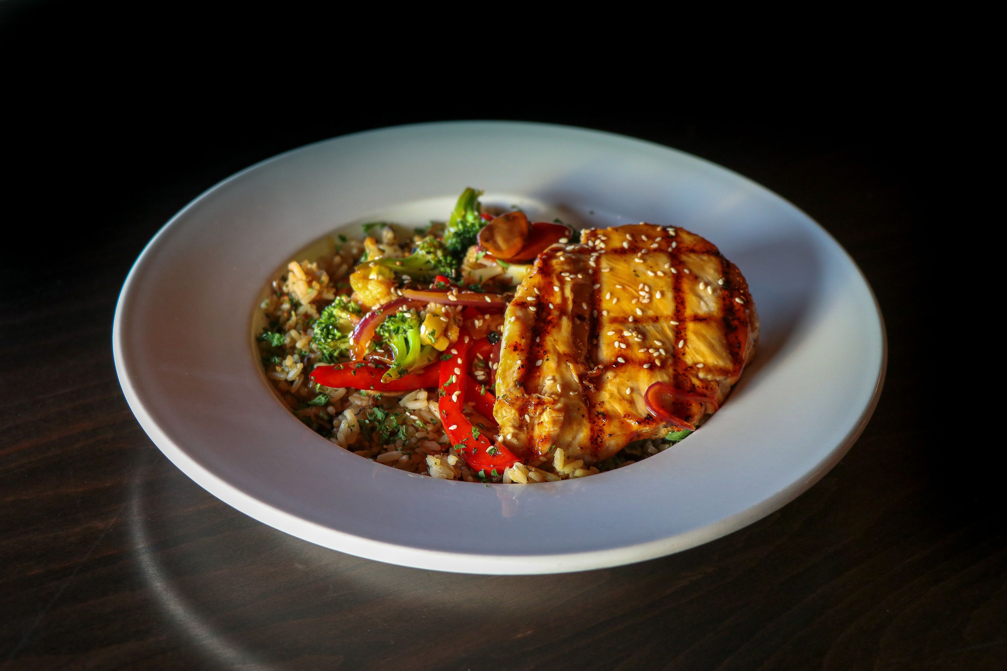A white plate with a grilled chicken breast, red bell peppers, broccoli, and a grain-based side dish.