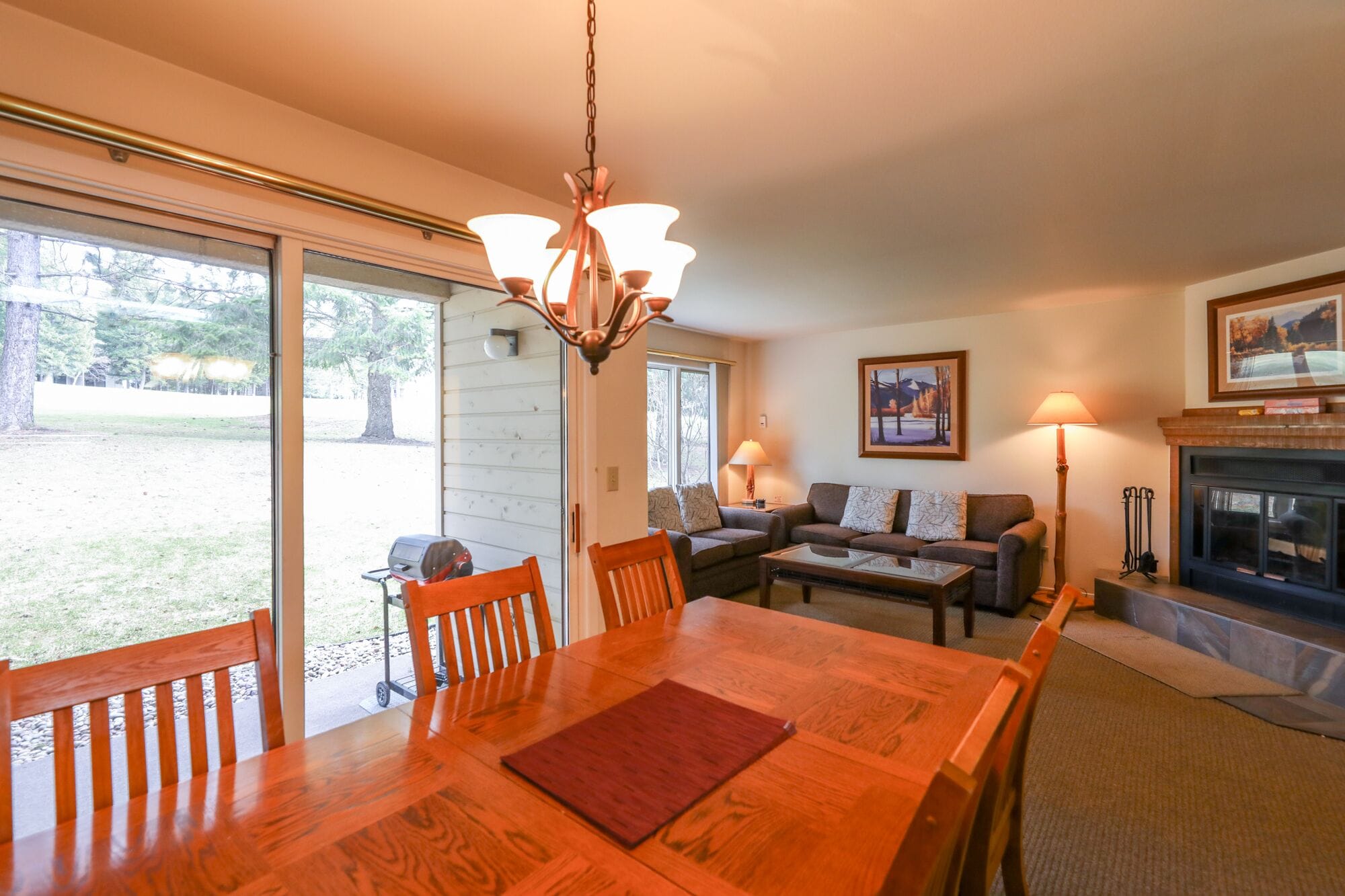 A dining area with a wooden table and chairs, a chandelier, a fireplace, and a view of the outdoors.