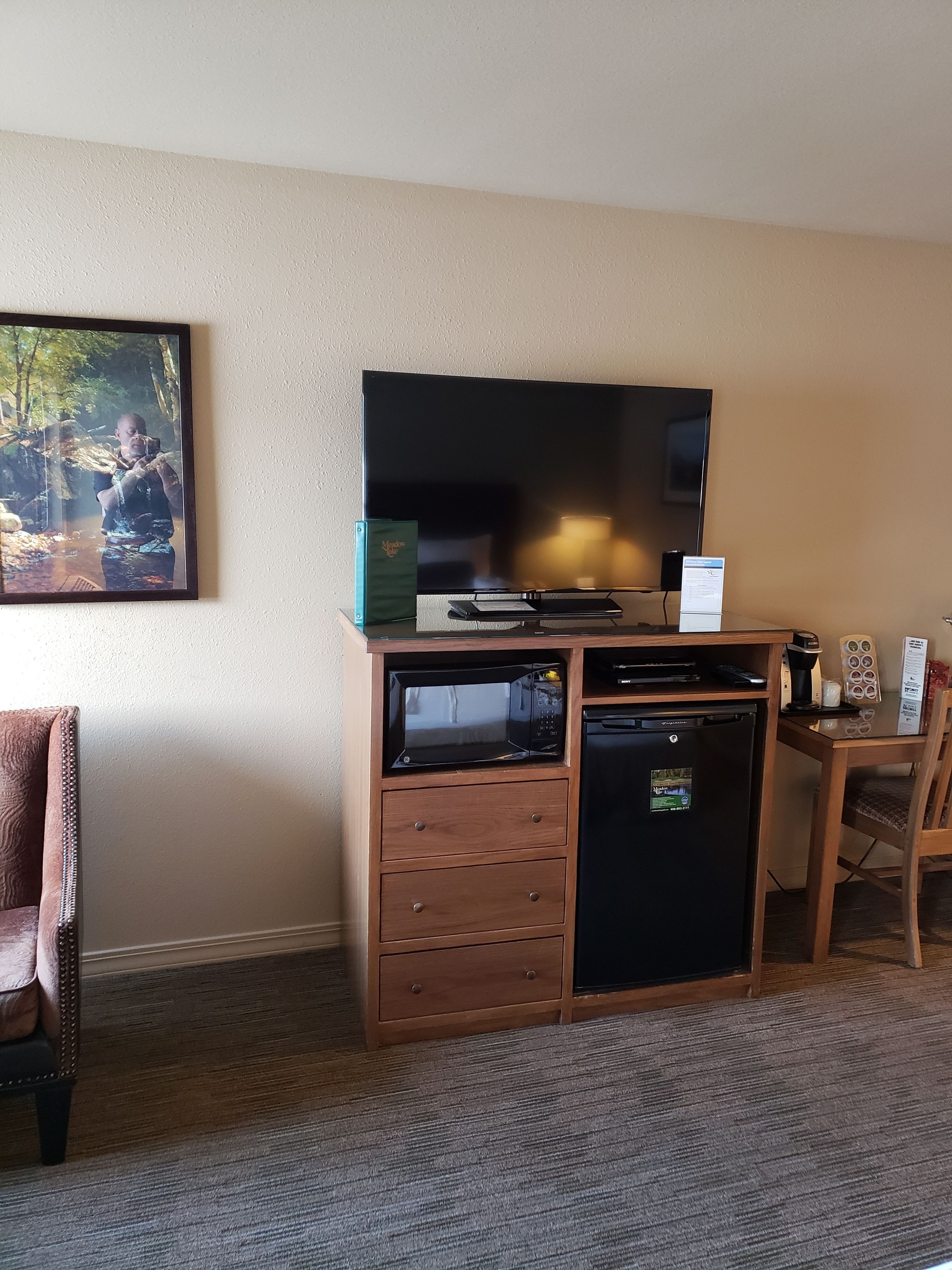 Habitación de hotel con televisión de pantalla plana, microondas, refrigerador pequeño, aparador con cajones, una silla y un cuadro en la pared.