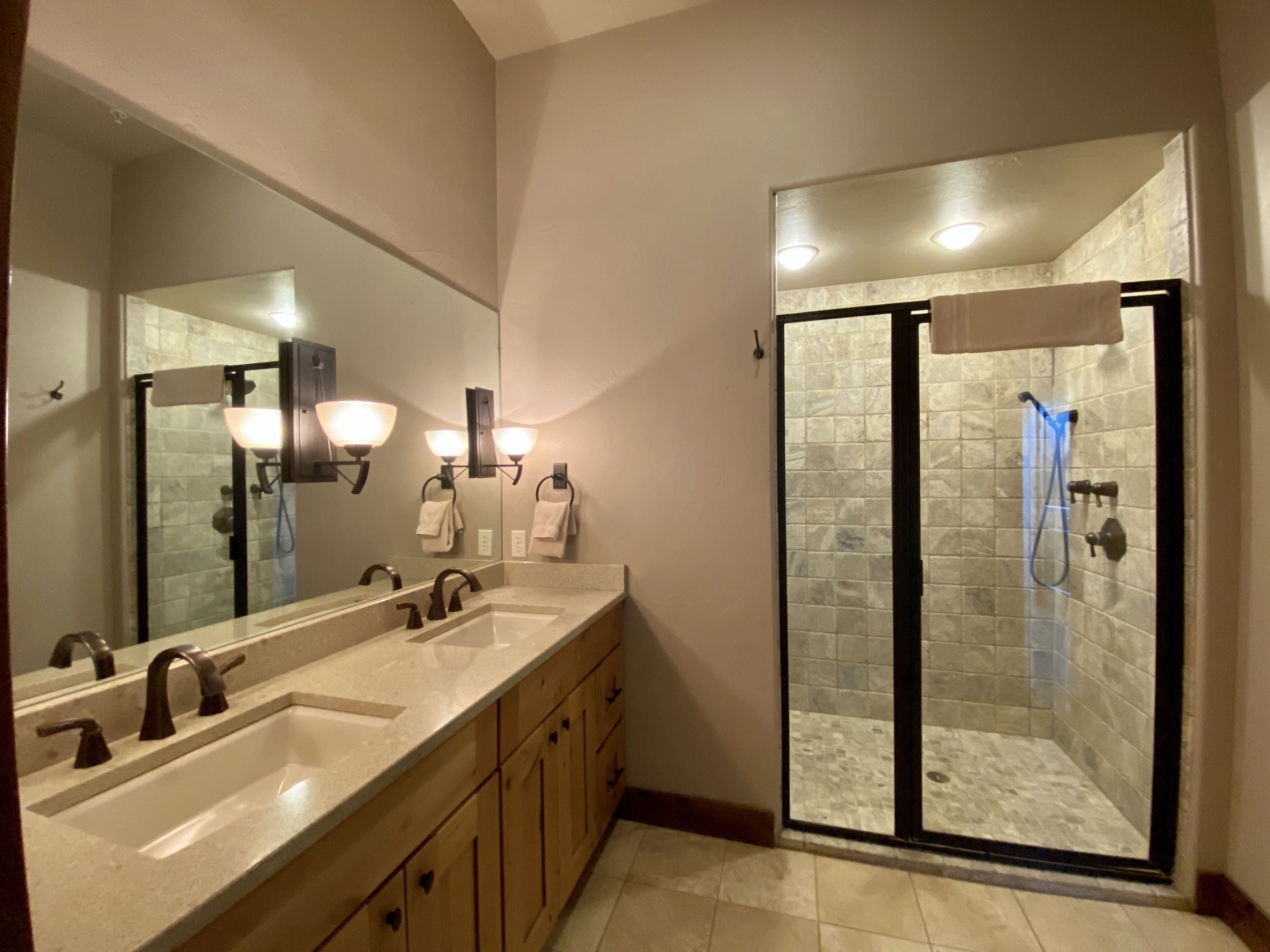 A bathroom with a large mirror, two sinks, a shower with glass doors, and wall-mounted lighting.