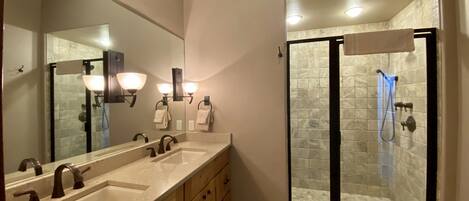 A bathroom with a large mirror, two sinks, a shower with glass doors, and wall-mounted lighting.