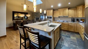 A modern kitchen with a central island, bar stools, wooden cabinets, a stone accent wall, and pendant lighting. - Meadow Lake Resort & Condos (Columbia Falls)