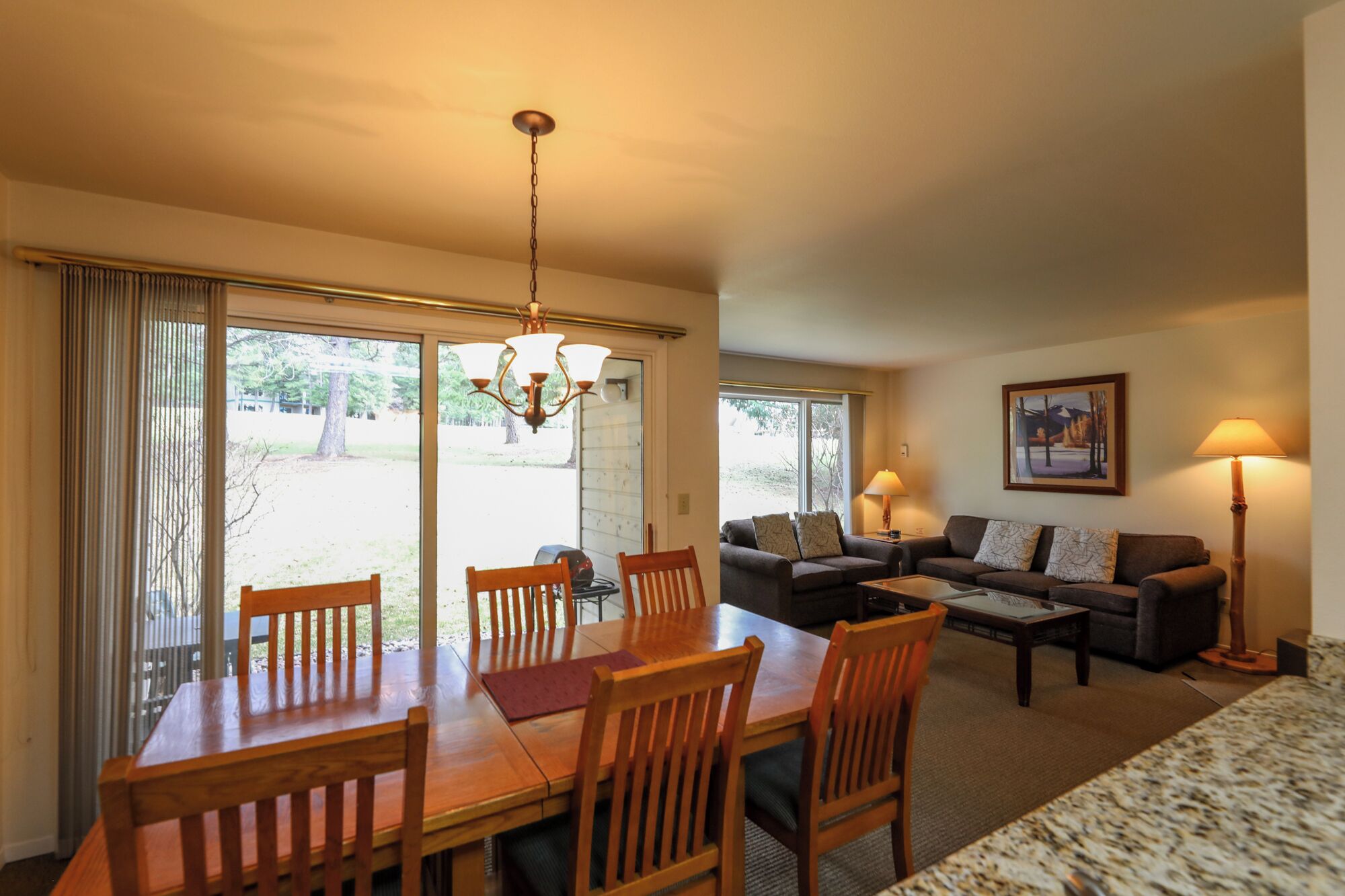 A spacious living area with a wooden dining table and chairs, a sofa, and a coffee table. There is a chandelier hanging from the ceiling, and a painting on the wall.
