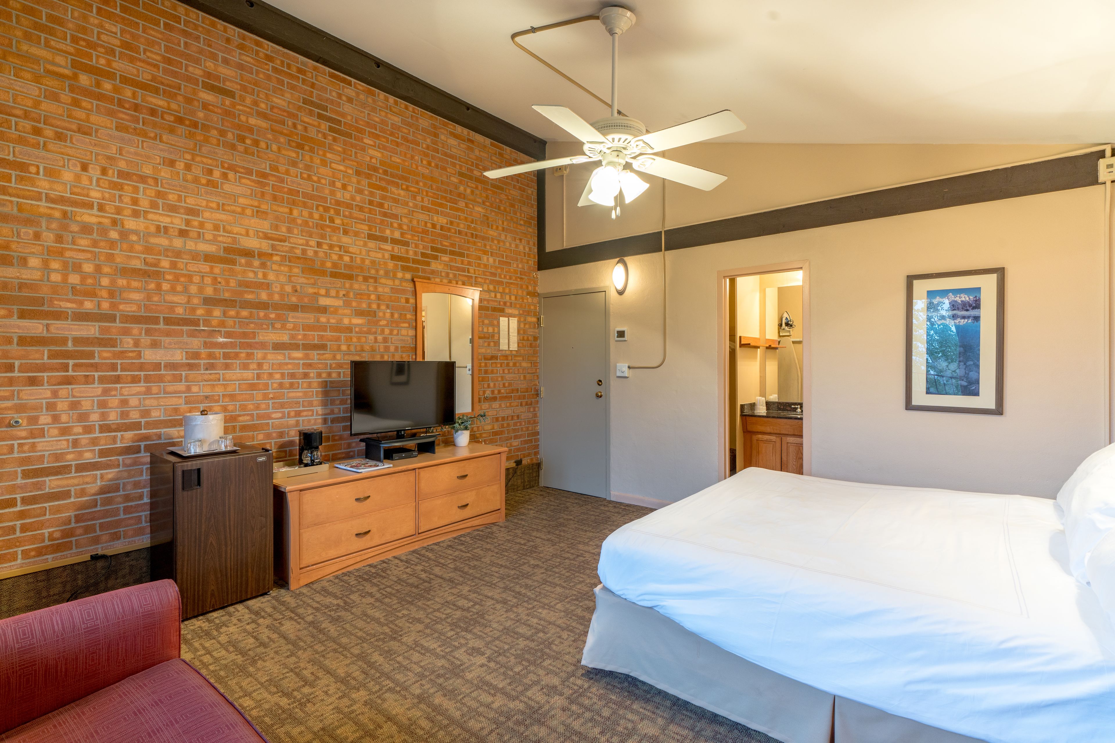 A hotel room with a brick wall, a bed, a desk with a TV, a dresser, and a ceiling fan.