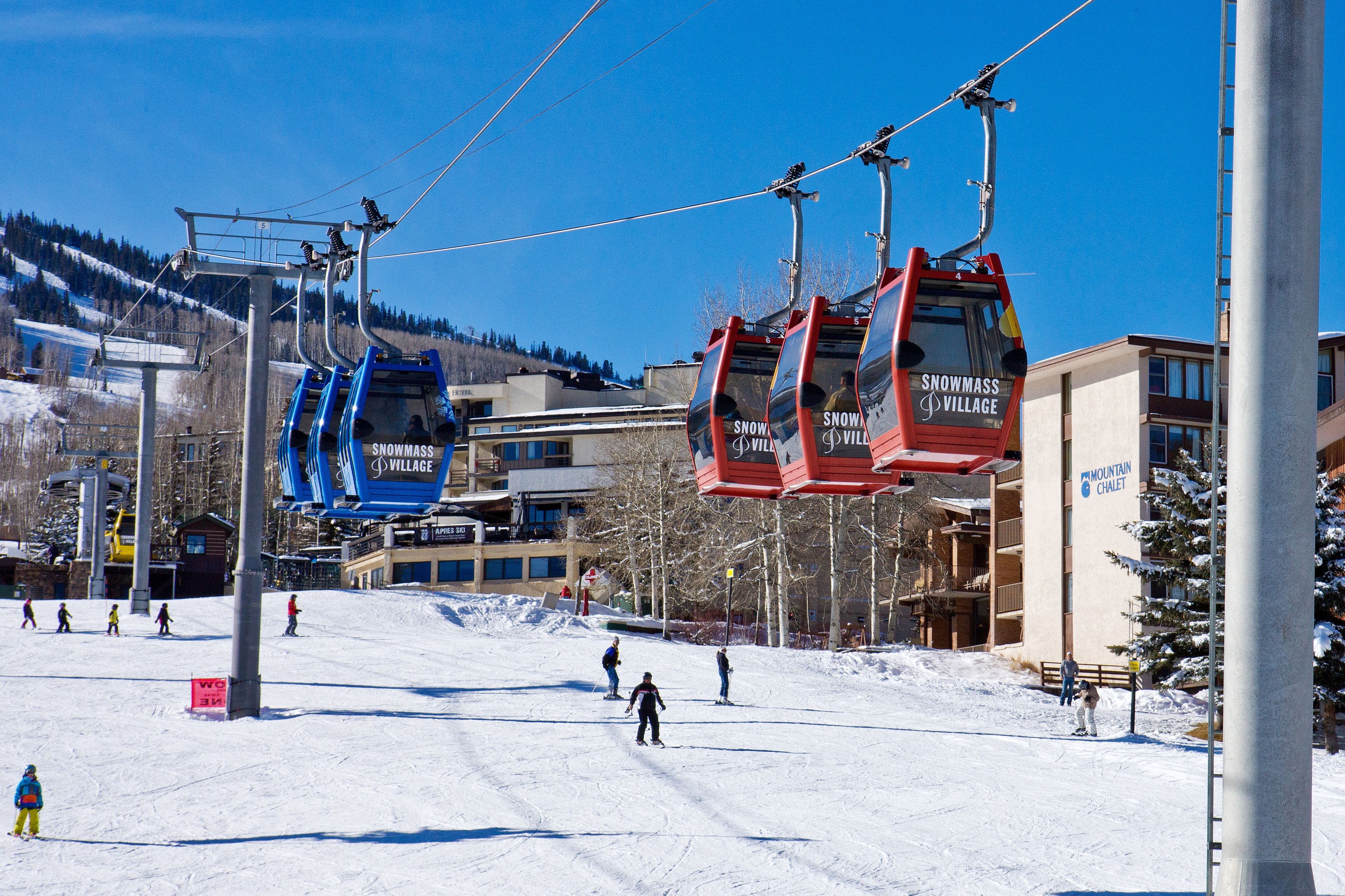 Un telesilla con cabinas identificadas como "Snowmass Village" y "Mountain Chalet" frente a un paisaje nevado y edificios.