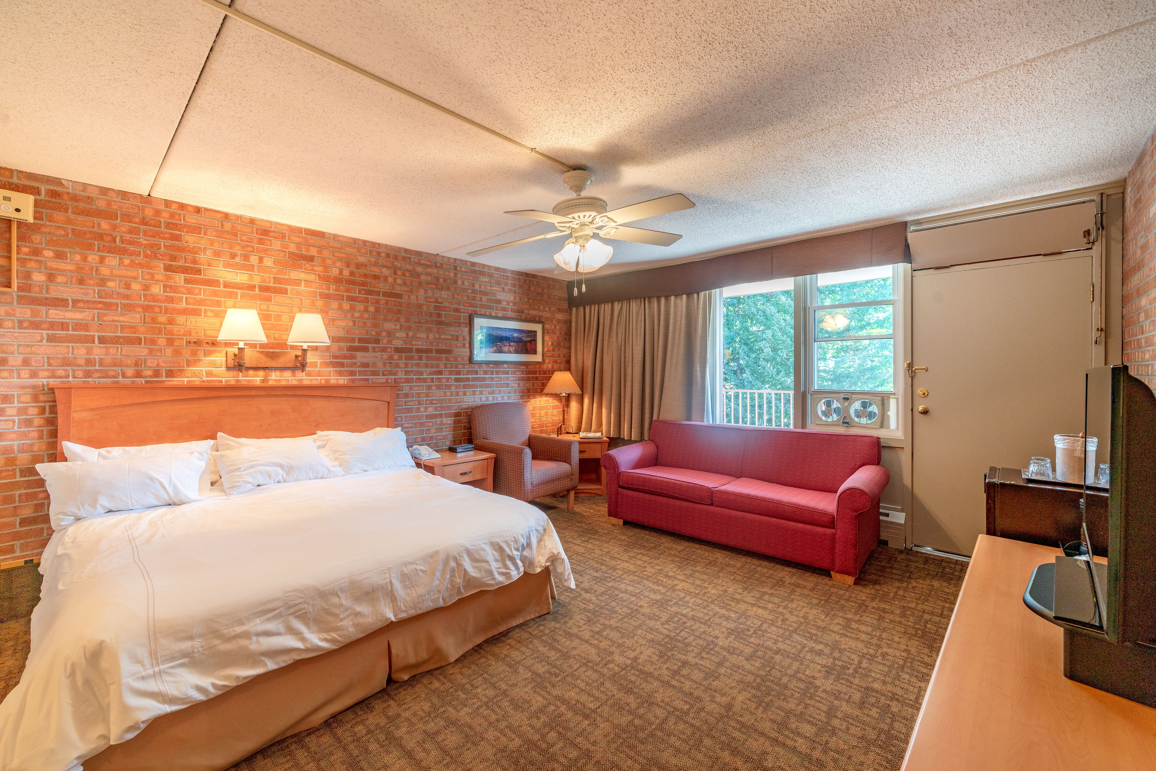 A hotel room with a large bed, a red sofa, a desk, a ceiling fan, and a window with curtains.