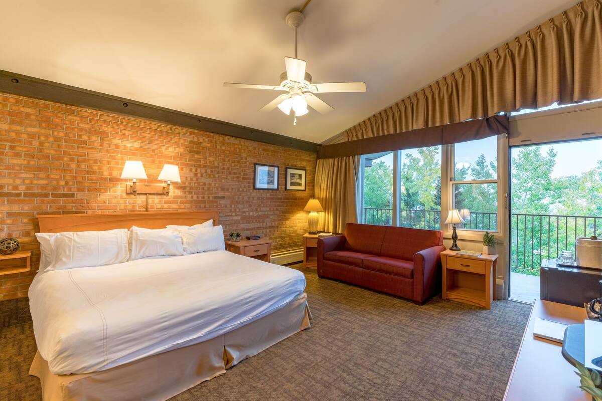 Une chambre d’hôtel avec un grand lit, un canapé rouge, un bureau, un ventilateur de plafond et une fenêtre donnant sur des arbres.