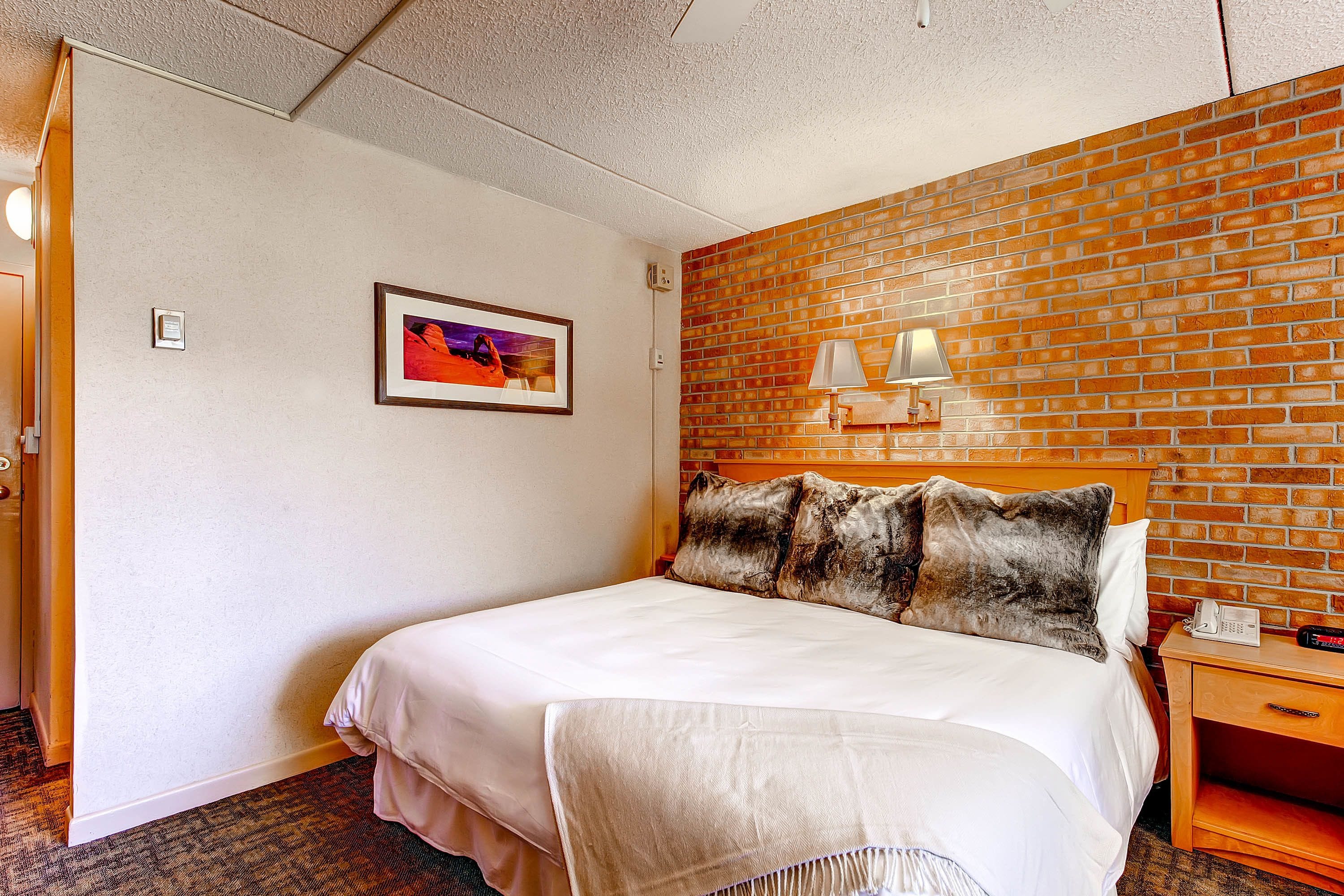 Quarto de hotel com uma cama, parede de tijolos decorativa, quadro emoldurado, mesa de cabeceira com abajur e telefone.