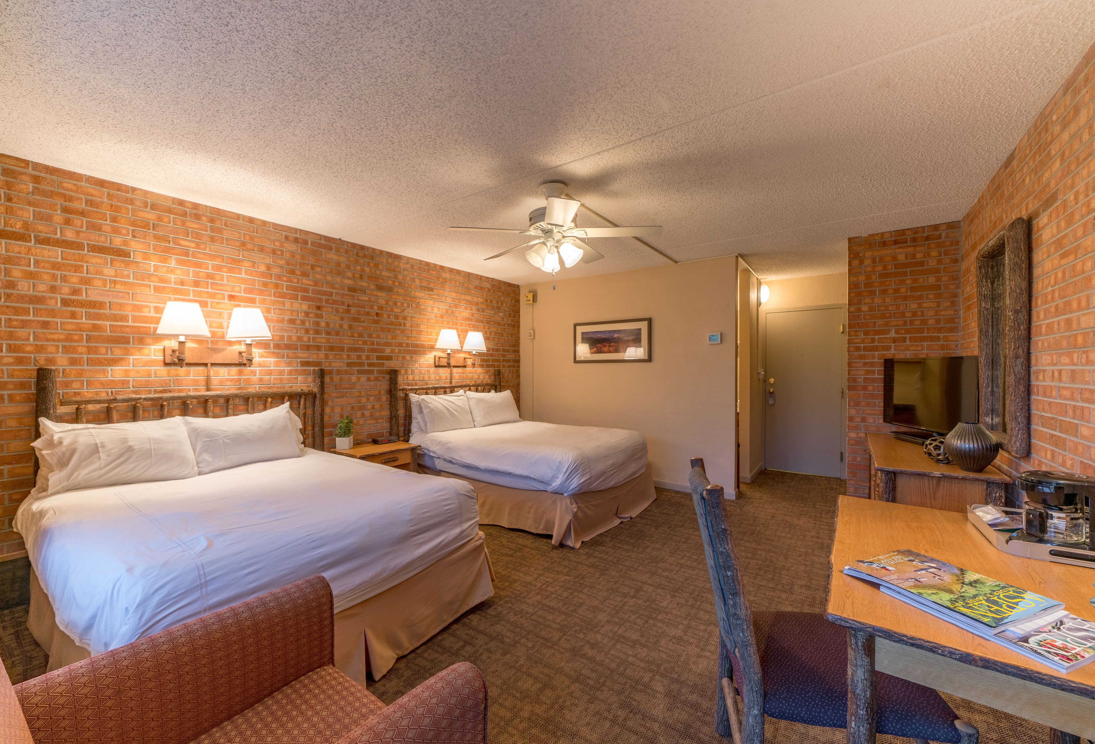 Quarto de hotel com duas camas, um ventilador de teto, parede de tijolos, escrivaninha de madeira e uma cadeira.