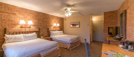 Habitación de hotel con dos camas, ventilador de techo, pared de ladrillo, escritorio de madera y una silla.