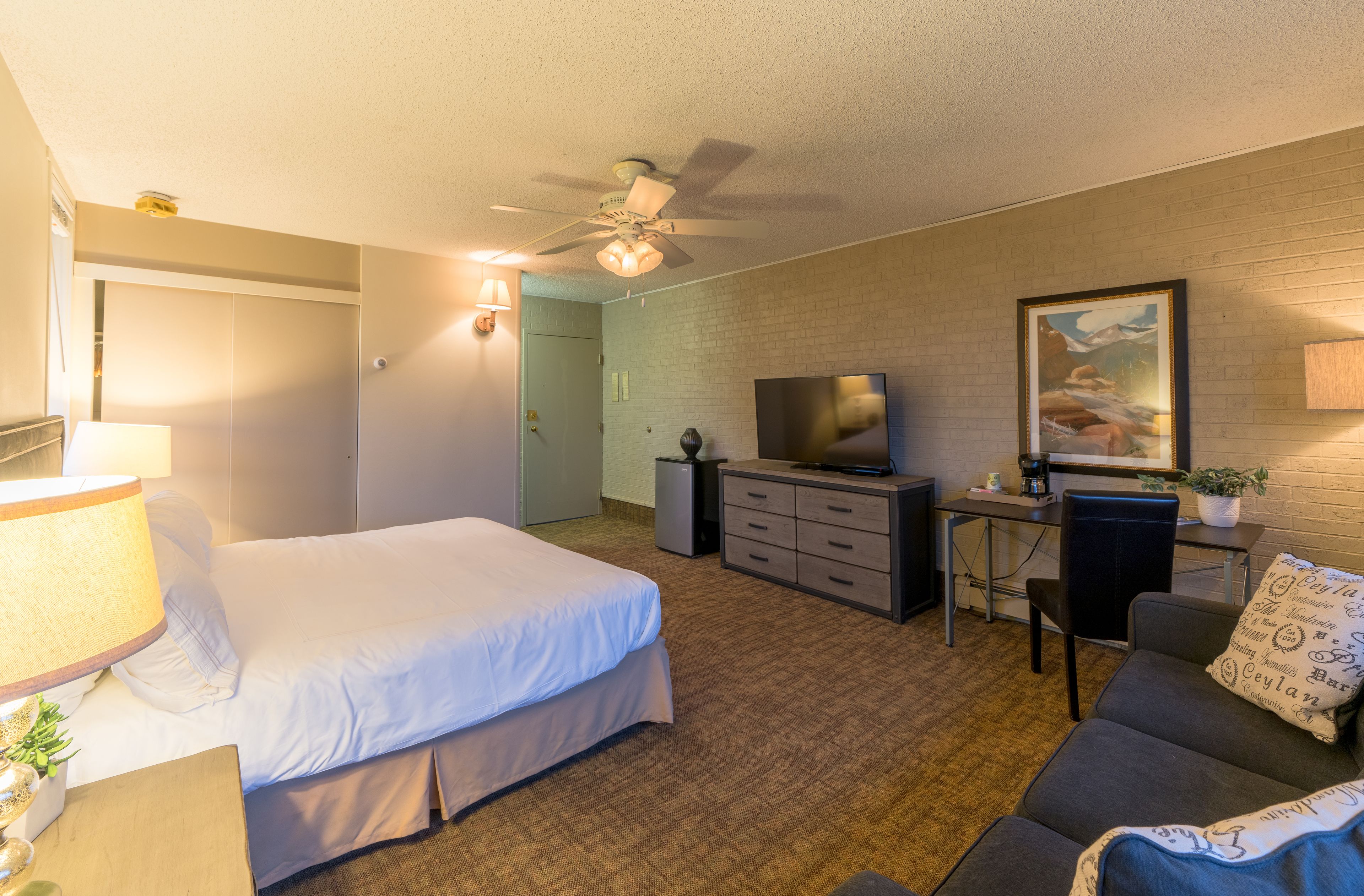 Une chambre d’hôtel avec un lit, un bureau avec une télévision, un ventilateur de plafond et un tableau au mur.