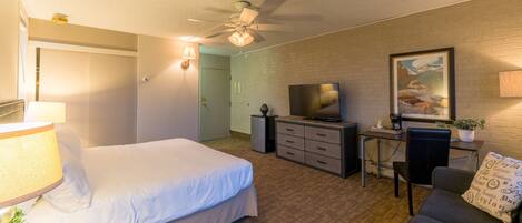 Habitación de hotel con cama, escritorio con televisor, ventilador de techo y un cuadro en la pared.