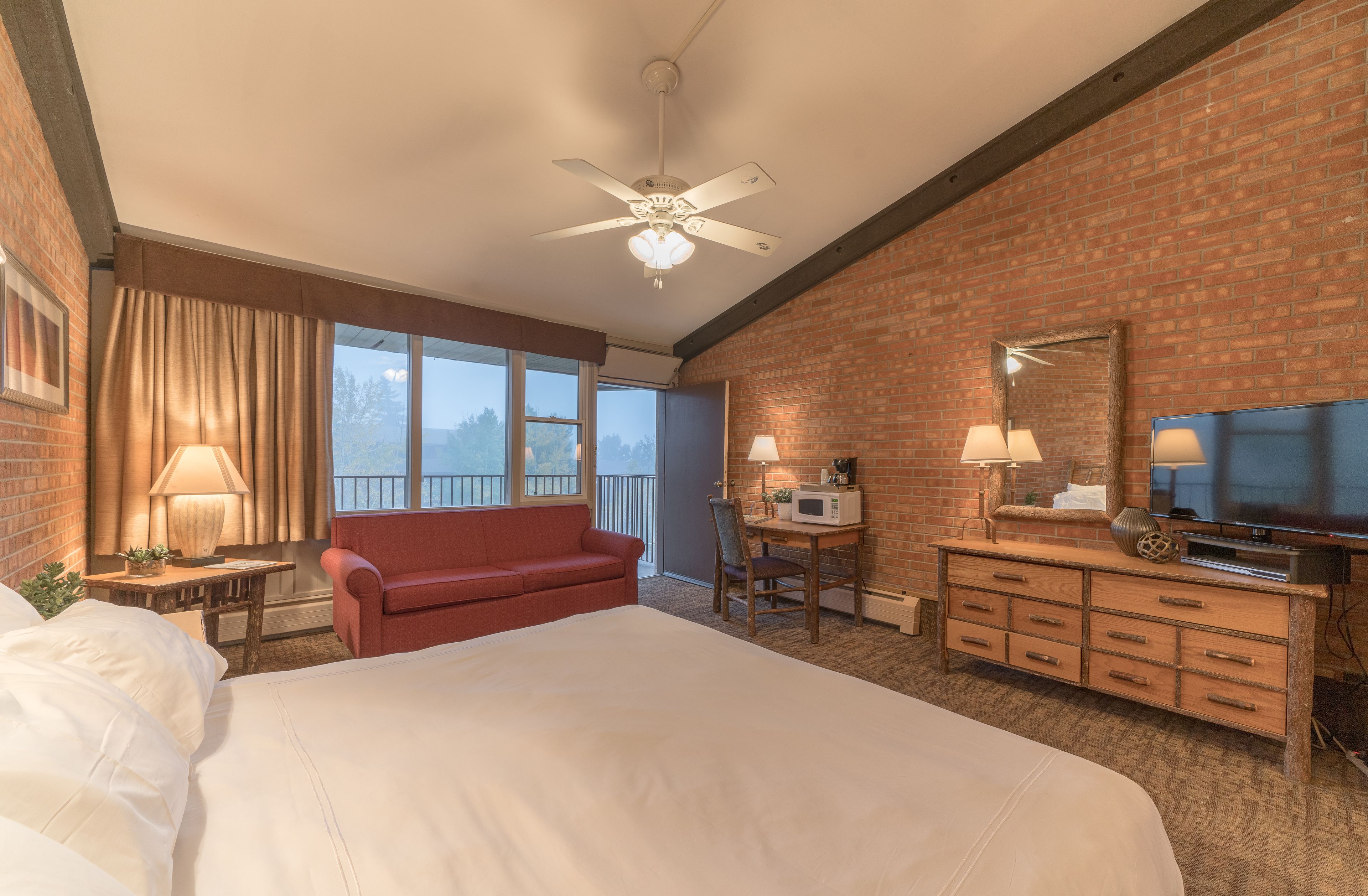 A hotel room with a large bed, a red sofa, a desk with a computer, a television, and a ceiling fan.