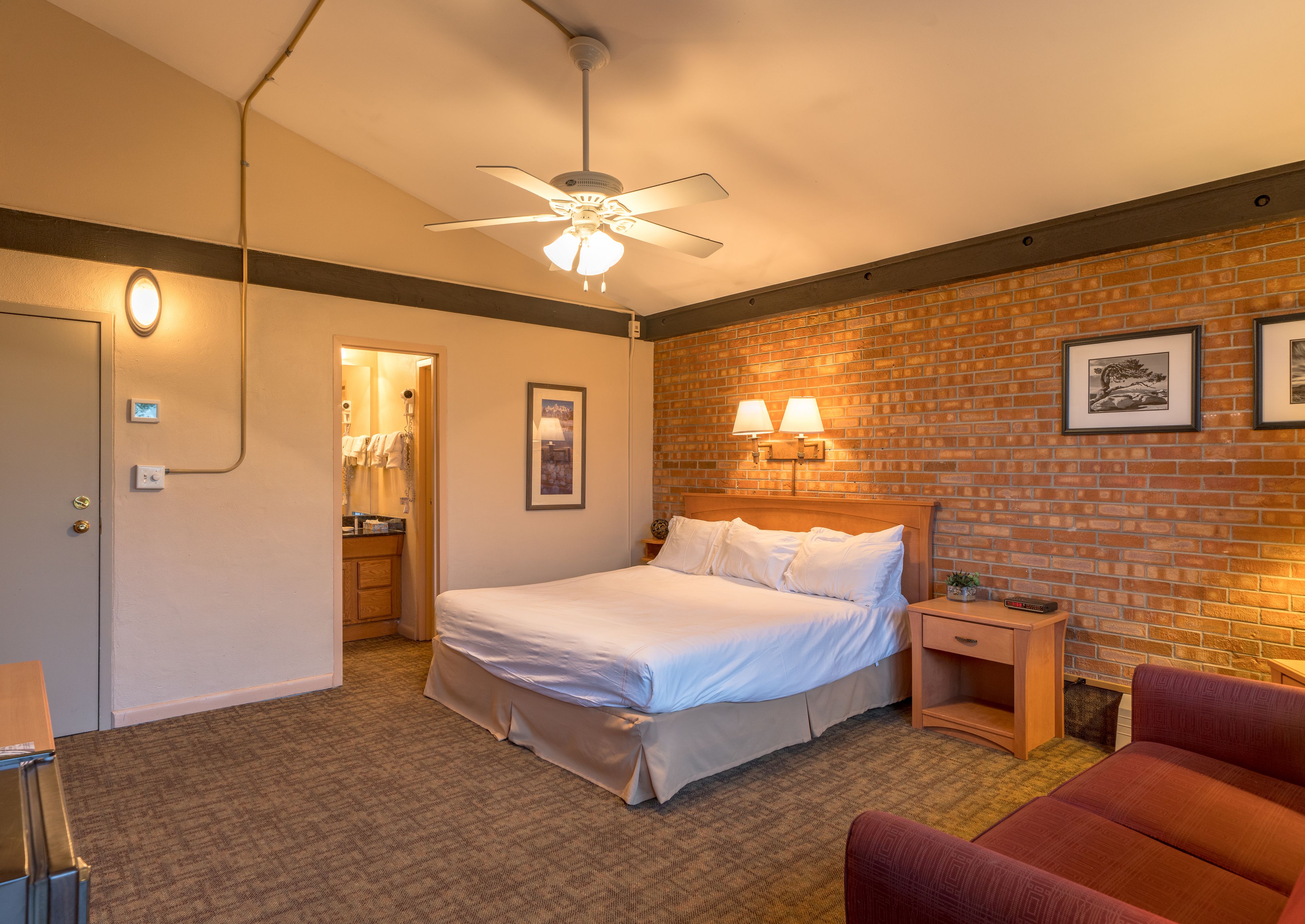 A hotel room with a bed, a wooden headboard, a brick wall, two wall-mounted lamps, a ceiling fan, and a red chair.