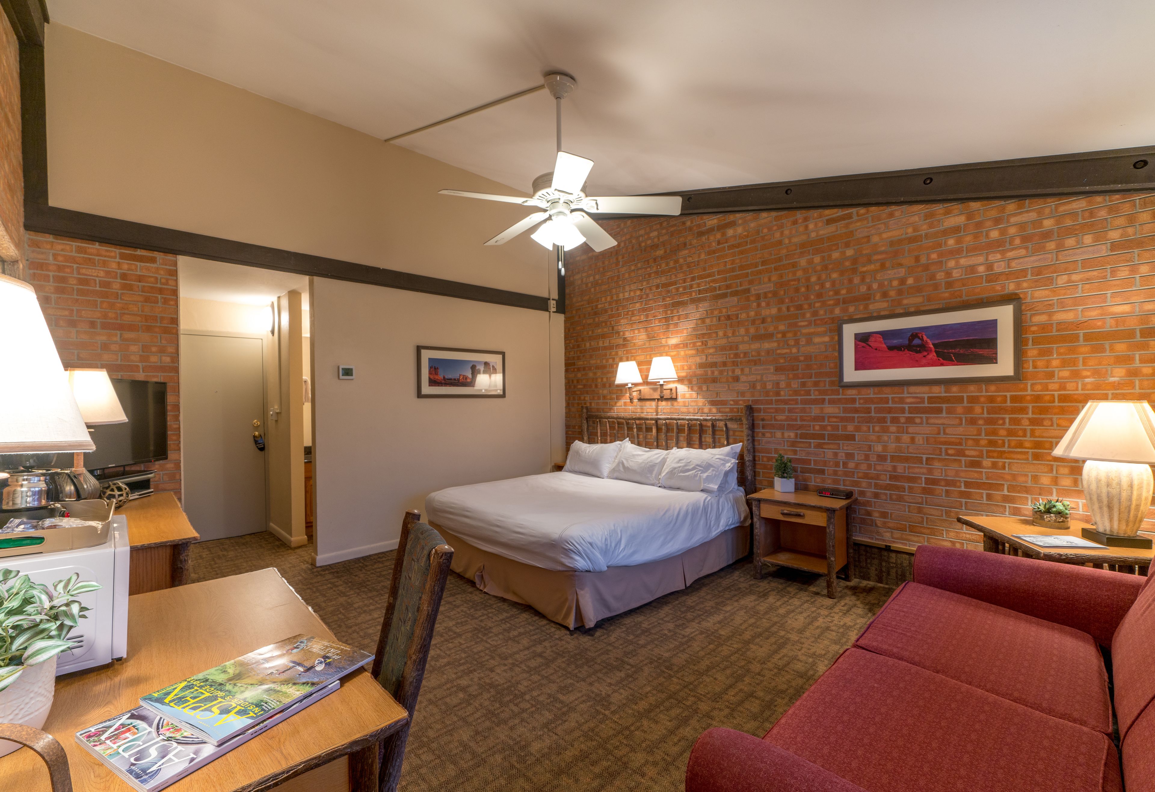 Ein Hotelzimmer mit einem Bett, einem roten Sofa, einem Schreibtisch mit Lampe, einem kleinen Tisch mit Pflanze und einem Deckenventilator.