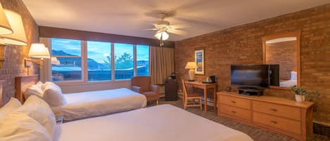 A hotel room with two beds, a desk, a television, and a large window with a mountain view.