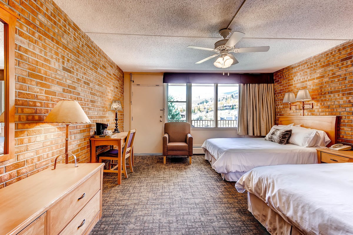Une chambre d’hôtel avec deux lits, un bureau, une chaise, une commode, un ventilateur de plafond et une fenêtre avec des rideaux.