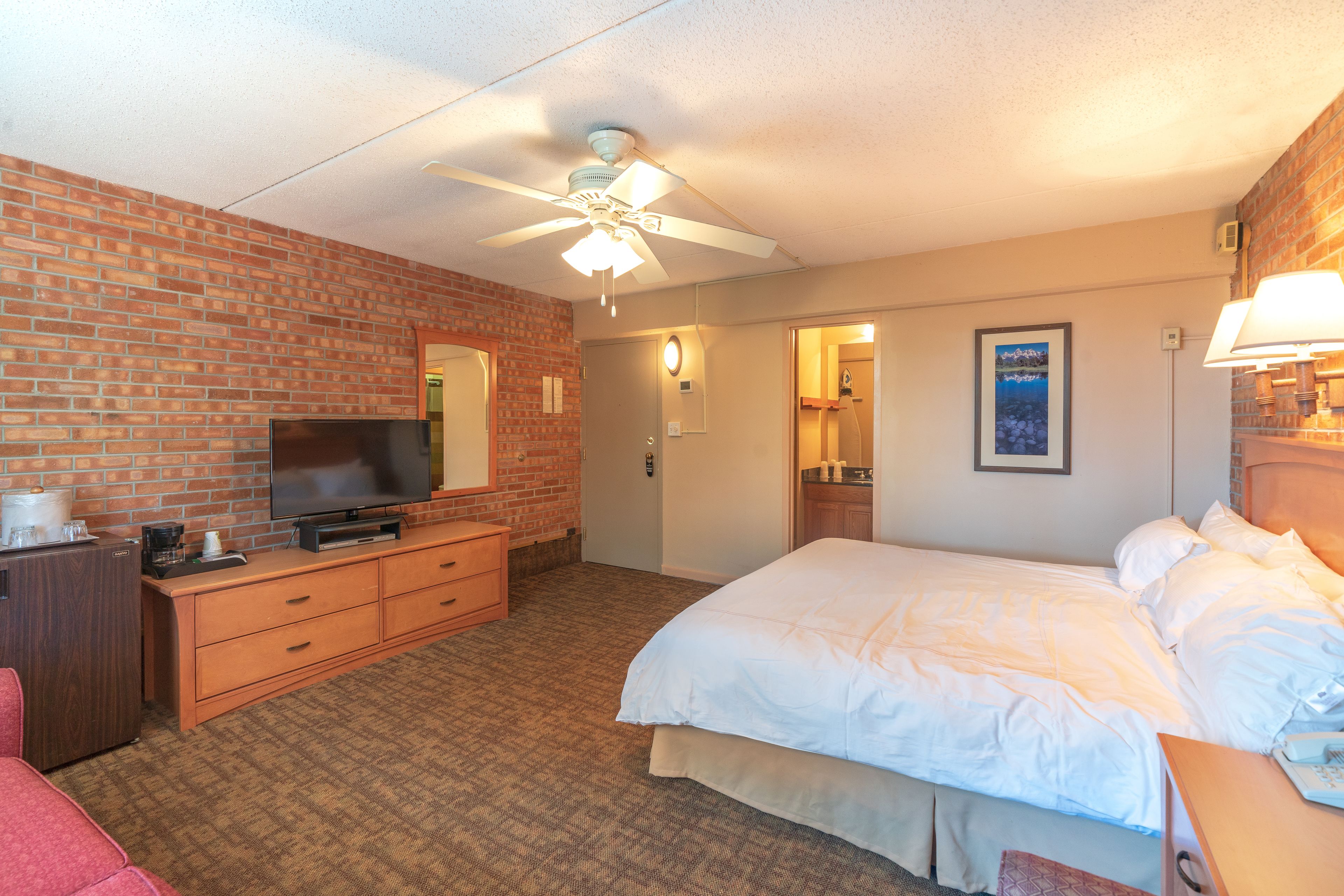 Une chambre d’hôtel avec un mur en briques, un ventilateur de plafond, un téléviseur à écran plat, un lit avec une literie blanche, une commode en bois et une salle de bain visible à travers une porte ouverte.