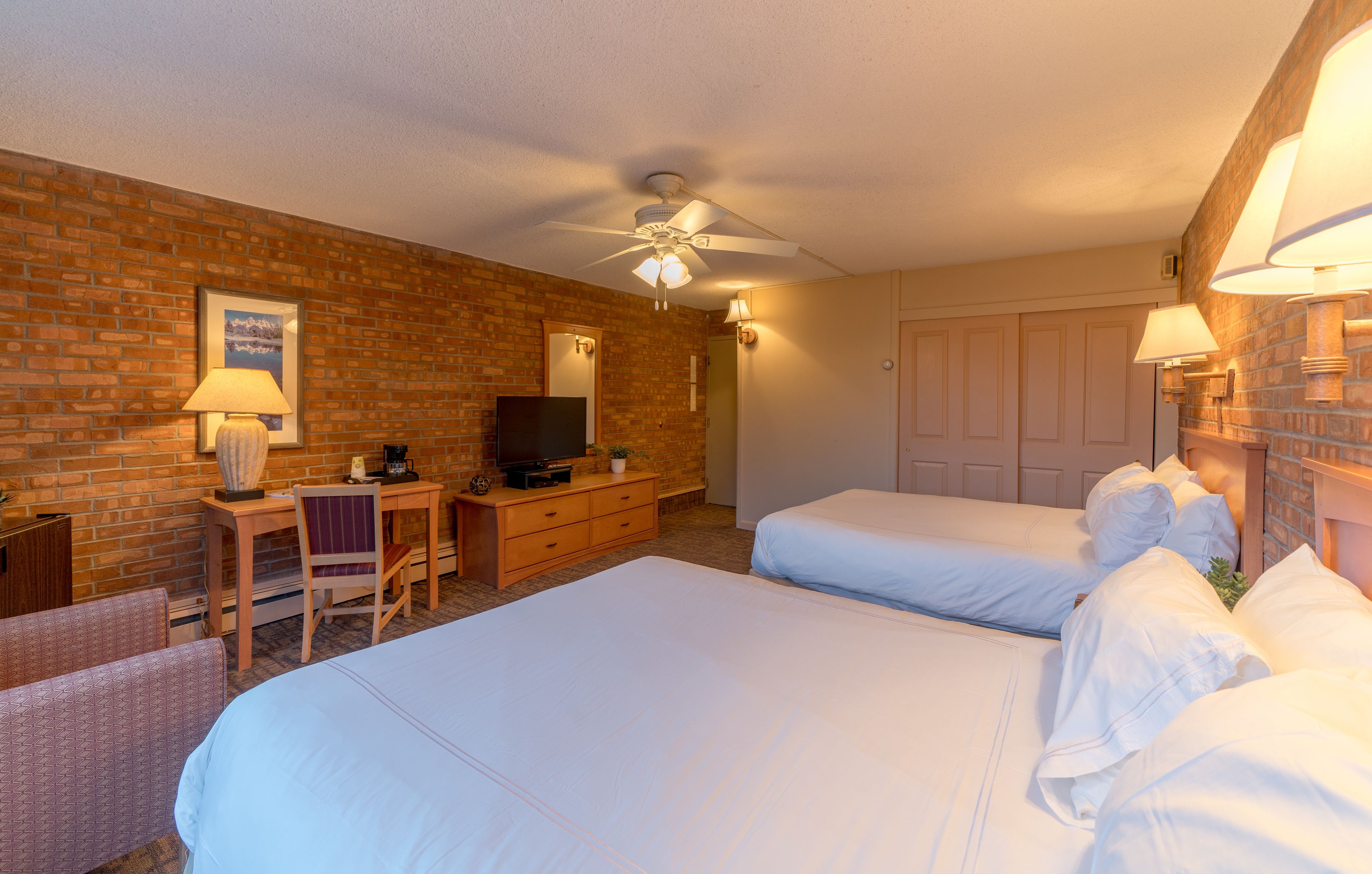 A hotel room with two beds, a desk with a chair, a television, and a brick wall.