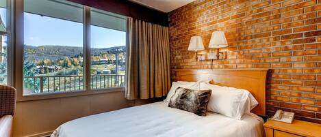 A hotel room with a large window, a brick wall, a bed with white linens, a nightstand with a lamp, and a view of a cityscape.