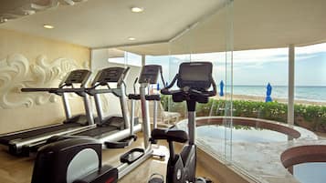 Une salle de sport équipée de tapis de course et d’un appareil elliptique, une grande fenêtre donnant sur la plage et un espace piscine.