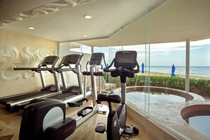 A gym with treadmills and an elliptical machine, a large window with a view of the beach, and a pool area. - Sunset Fishermen Beach Resort Playa del Carmen - All inclusive (Playa del Carmen)